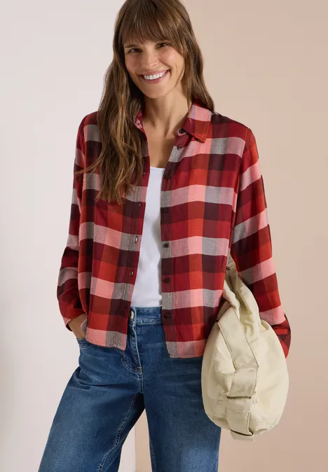 Une femme souriante porte une chemise à carreaux rouge, t-shirt blanc, jean bleu et tient un sac beige.