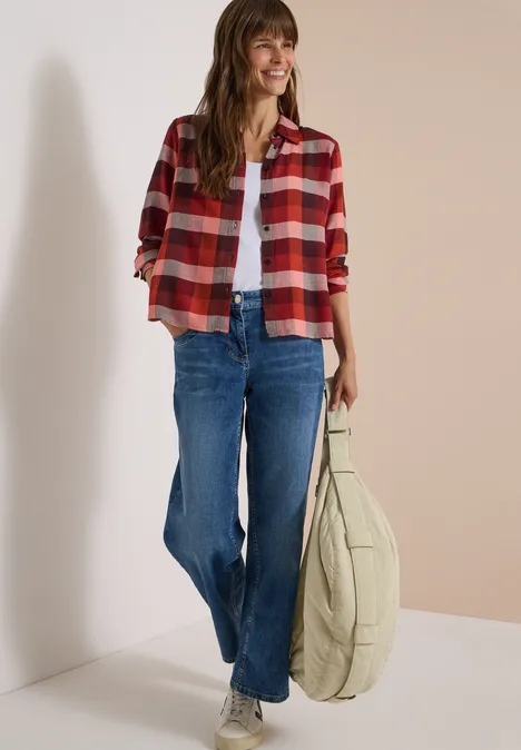 Jeune femme souriante en chemise à carreaux rouge, débardeur blanc, jean bleu et grand sac beige.