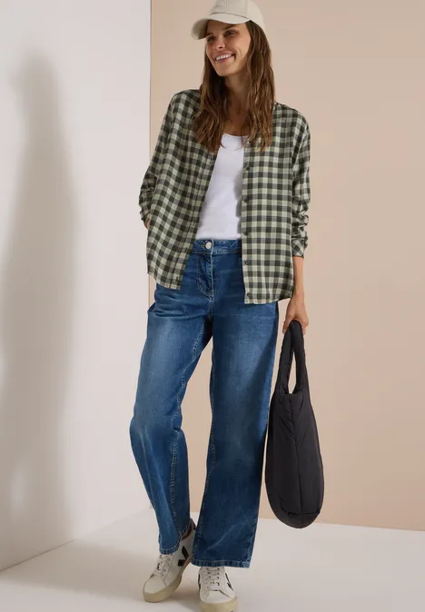 Overhemd met lange mouwen en ruitjespatroon khaki Frau in kariertem Hemd, weißem Top, weiten Jeans, Sneakers und Cap hält schwarze Tasche und lächelt vor beiger Wand.