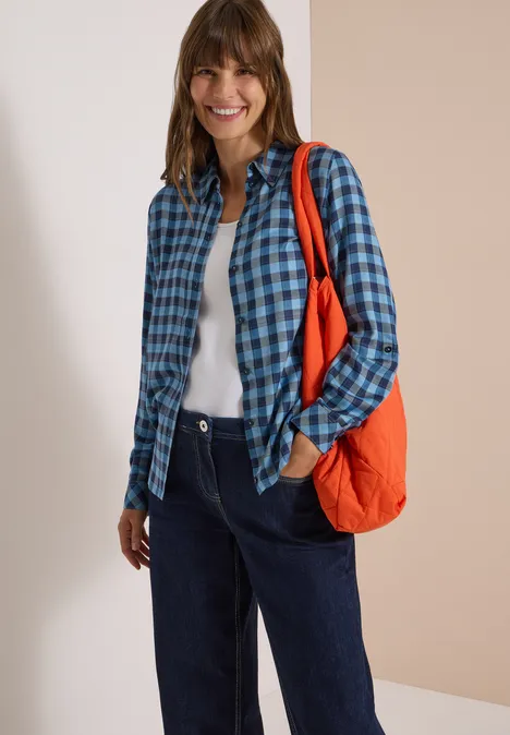Frau mit blau kariertem Hemd, weißem Top, dunklen Jeans und orangener Tasche, lächelnd.