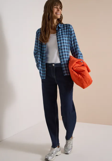 Frau in blauem Karohemd, weißem Top, dunklen Jeans und silbernen Sneakern hält orangefarbene Jacke.