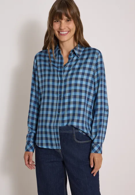 Frau in blau kariertem Hemd und Jeans vor weißem Hintergrund, lächelnd.