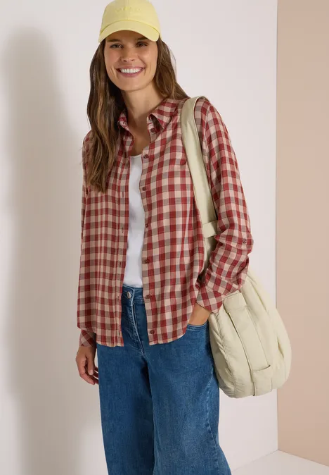 Lächelnde Frau in roter karierter Bluse, weißem T-Shirt, blauen Jeans, gelber Cap und beigem Beutel.