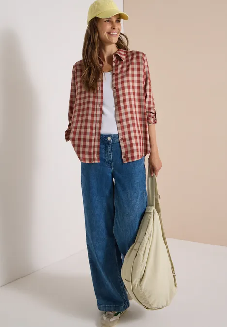 Junge Frau in weiten Jeans, weißem T-Shirt, rot kariertem Hemd, gelber Cap und grüner Tasche.