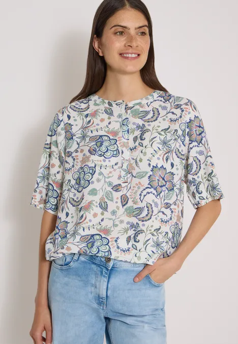 Frau in weißer Bluse mit pastellfarbenem Blumenmuster und lockerer Passform, kombiniert mit hellen Jeans.
