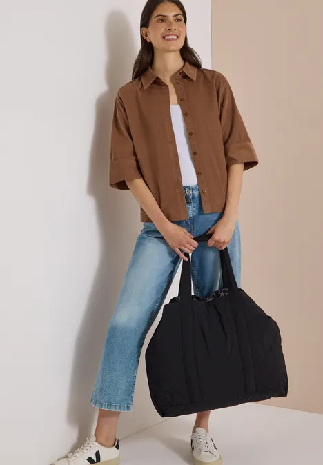 Chemisier à manches courtes uni creamy mocha Frau im Outfit mit Jeans, weißem Top, brauner Hemdbluse und weißen Sneakern, die eine große schwarze Tasche trägt.