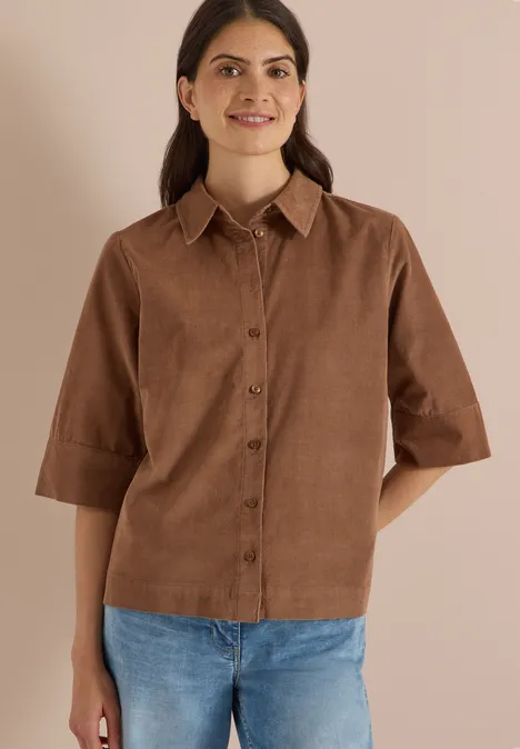 Chemisier à manches courtes uni creamy mocha Eine Frau trägt eine braune Cord-Bluse mit 3/4-Ärmeln, Knopfleiste und Kragen zu hellen Jeans vor beigem Hintergrund.