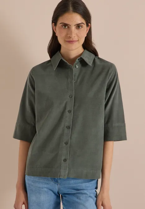 Chemisier à manches courtes uni dried olive green Frau im dunkelgrünen, knöpfbaren Hemd mit Dreiviertelärmeln und blauen Jeans vor neutralem Hintergrund.