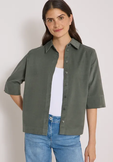 Chemisier à manches courtes uni dried olive green Frau im grünen Hemd mit halblangen Ärmeln, weißem Top und blauen Jeans, leicht lächelnd.