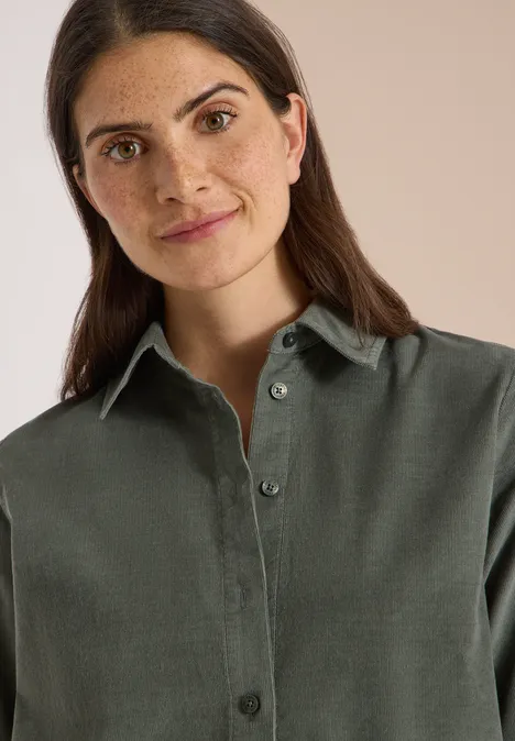 Chemisier à manches courtes uni dried olive green Porträt einer lächelnden Frau mit dunklem Haar in grünem Hemd vor neutralem Hintergrund.