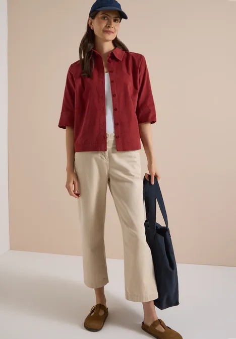 Chemisier à manches courtes uni russet red Eine Frau trägt rote Halbarmbluse, weißes T-Shirt, beige Hose, braune Schuhe, blaue Tasche und Cap.