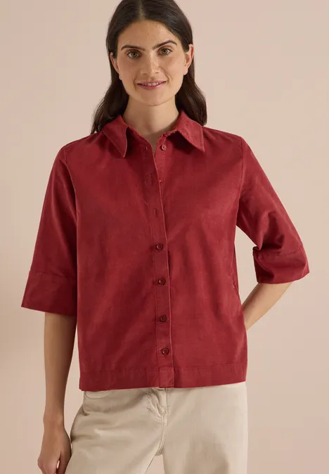 Chemisier à manches courtes uni russet red Frau trägt rote Cordbluse mit halblangen Ärmeln und beige Hose vor hellem Hintergrund.