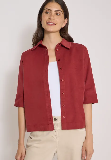 Chemisier à manches courtes uni russet red Frau in bordeauxrotem Hemd mit 3/4-Ärmeln, weißem Top und heller Hose vor neutralem Hintergrund.