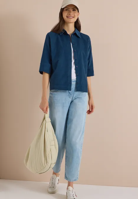 Chemisier à manches courtes uni moonlight blue Eine lächelnde Frau trägt Jeansjacke, Jeans, weiße Sneakers, Baseballcap und hält eine große beige Tasche.