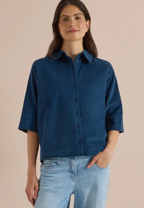 Chemisier à manches courtes uni moonlight blue Eine Frau in blauem Hemd mit kurzen Ärmeln und heller Jeans vor neutralem Hintergrund.