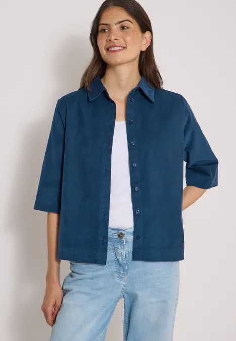 Chemisier à manches courtes uni moonlight blue Frau trägt blaues Jackett über weißem Top und Jeans, steht vor neutralem Hintergrund.