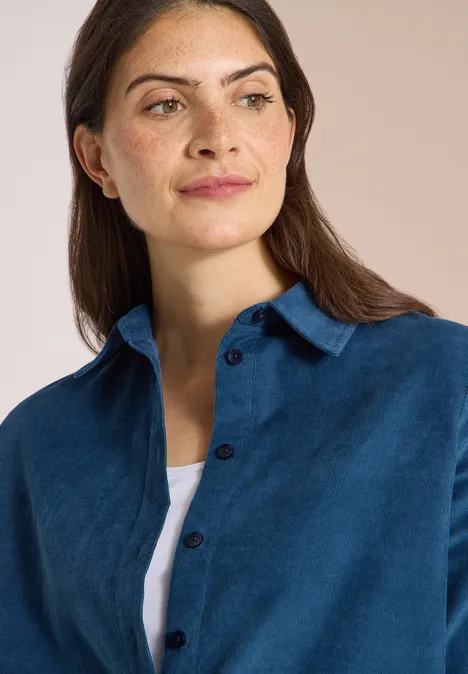 Chemisier à manches courtes uni moonlight blue Eine Frau mit braunen Haaren trägt ein blaues Cordhemd über einem weißen Top vor hellem Hintergrund.