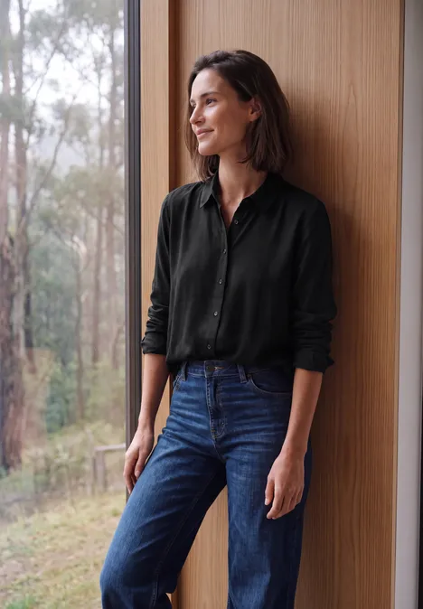 Eine lächelnde Frau in schwarzem Hemd und Jeans lehnt an einer Holzwand mit Blick ins Grüne.