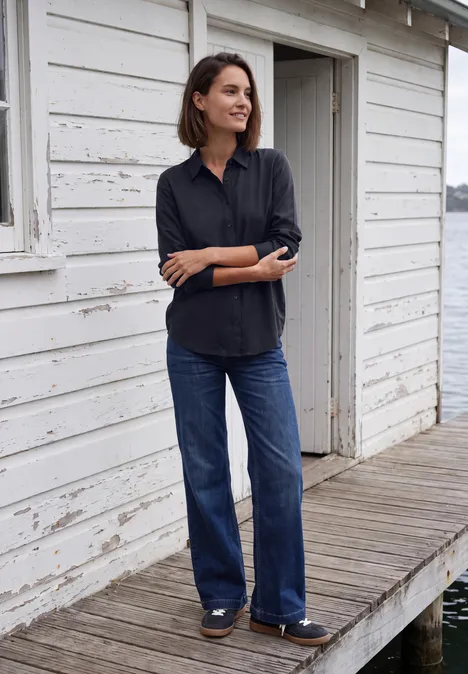 Een glimlachende vrouw in blouse en jeans voor een wit huis bij het water.
