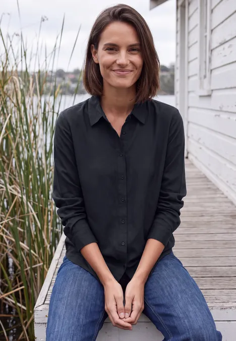 Een glimlachende vrouw in een zwarte blouse en spijkerbroek zit op een houten vlonder bij het water en riet.