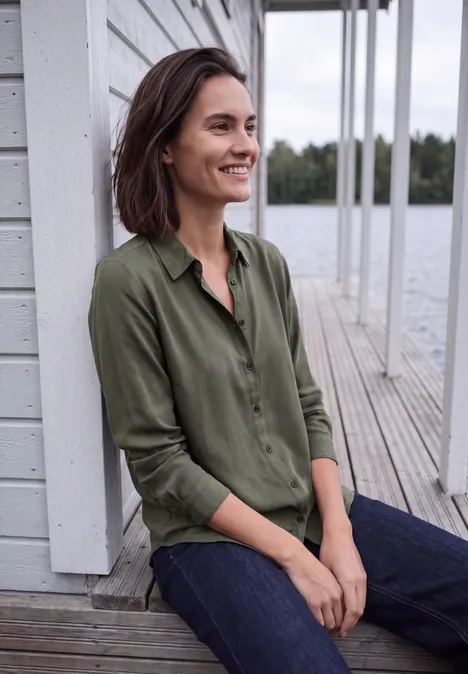 Eine lächelnde Frau in grüner Bluse sitzt entspannt auf einem Holzsteg am Wasser.