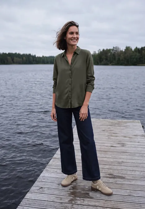Eine Frau steht auf einem Holzsteg am See, trägt grüne Bluse, blaue Jeans und helle Schuhe.
