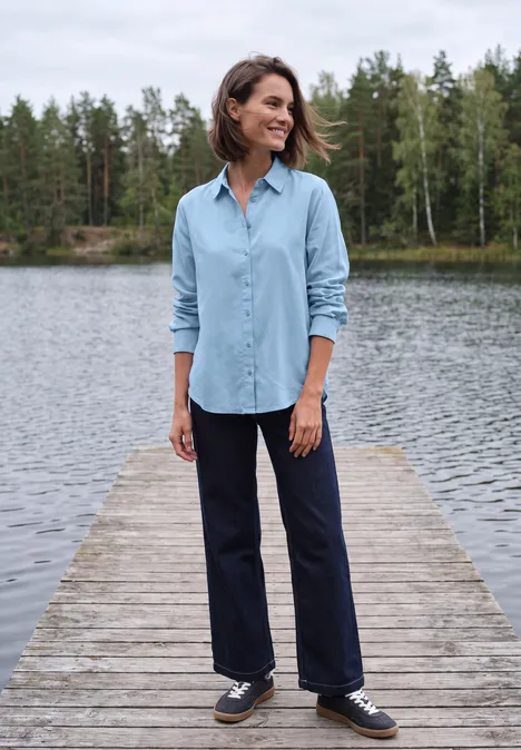 Langarm Hemdbluse mit Knopfleiste cosmopolitan blue Frau steht auf einem Steg an einem See, trägt hellblaues Hemd und dunkle Hose, lächelt, im Hintergrund Bäume.