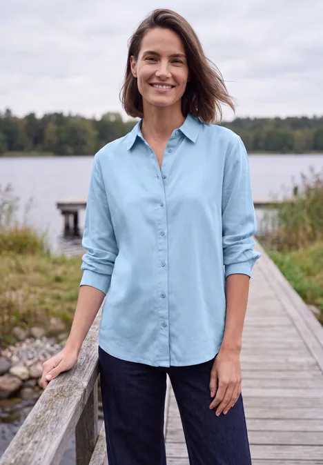 Langarm Hemdbluse mit Knopfleiste cosmopolitan blue Frau mit blauem Hemd steht lächelnd auf einem Steg an einem See, umgeben von Wiesen und Bäumen.
