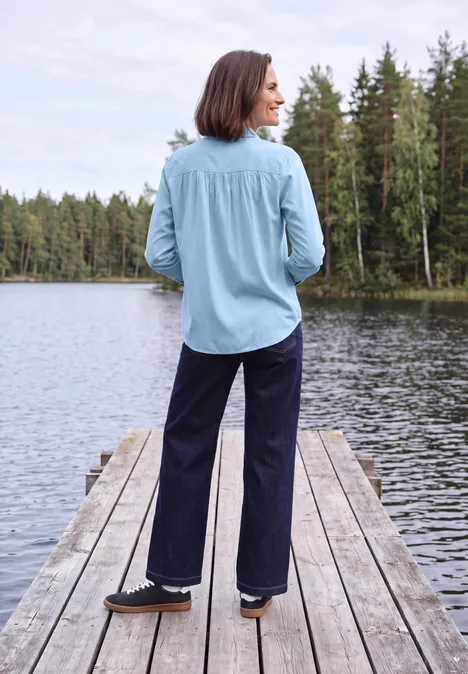 Langarm Hemdbluse mit Knopfleiste cosmopolitan blue Eine Frau steht in hellblauem Hemd und dunklen Hosen auf einem Steg am See, umgeben von Wäldern.