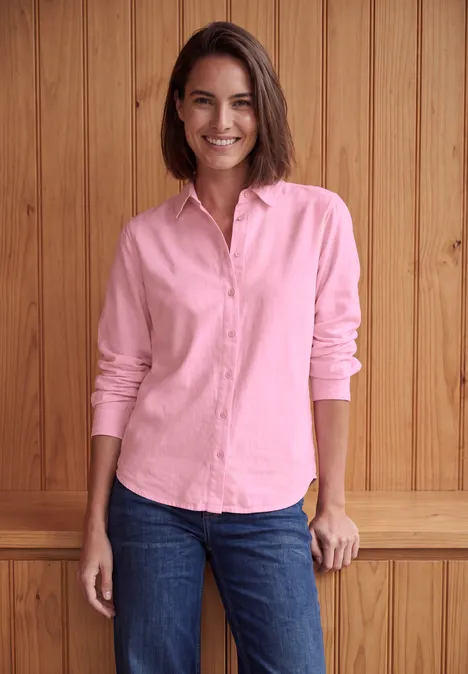 Vrouw met roze blouse en spijkerbroek lacht en staat voor een houten wand.