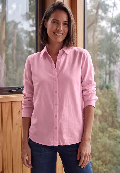 Een glimlachende vrouw in een roze blouse en spijkerbroek voor een raam met bomen.