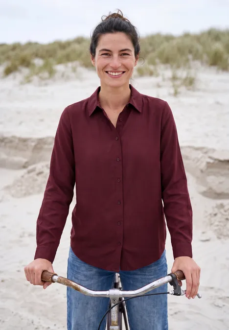 Chemisier à manches longues avec patte de boutonnage red wine Une femme souriante en chemise rouge et jean tient son vélo sur une plage de dunes.