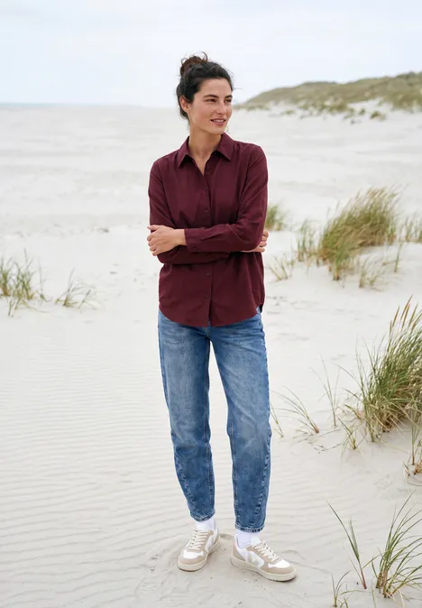 Chemisier à manches longues avec patte de boutonnage red wine Une femme en jean et chemise sourit debout sur une plage de dunes avec herbe, ciel clair.