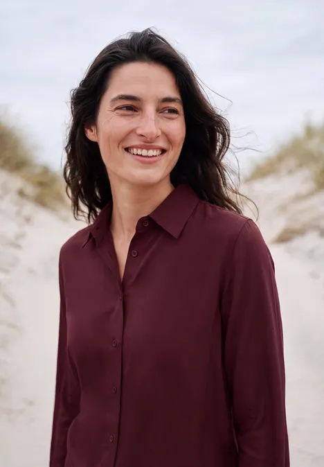 Chemisier à manches longues avec patte de boutonnage red wine Une femme sourit sur la plage devant les dunes.