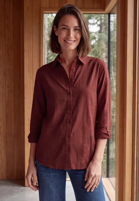 Eine lächelnde Frau in rotem Hemd und Jeans steht in einem modernen Holzhaus vor großen Fenstern.