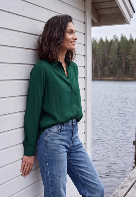 Eine Frau in grünem Hemd und Jeans lehnt an einer weißen Holzwand, im Hintergrund ein ruhiger See und Wald.