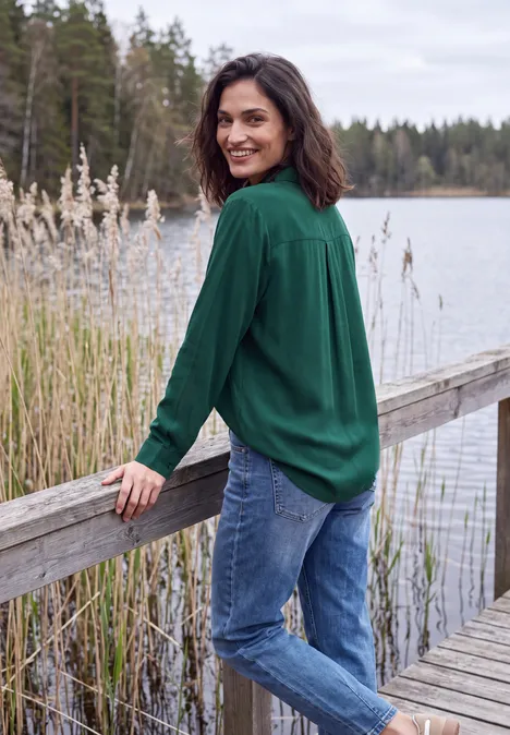 Frau im grünen Hemd lehnt lächelnd am Holzsteg an einem See, umgeben von Schilf und Wald im Hintergrund.