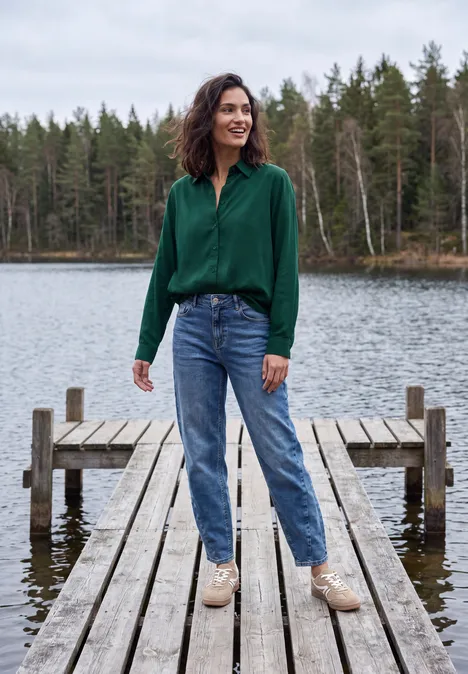 Frau in grüner Bluse, Jeans und hellen Schuhen steht lächelnd auf einem Holzsteg am See, Wald im Hintergrund.