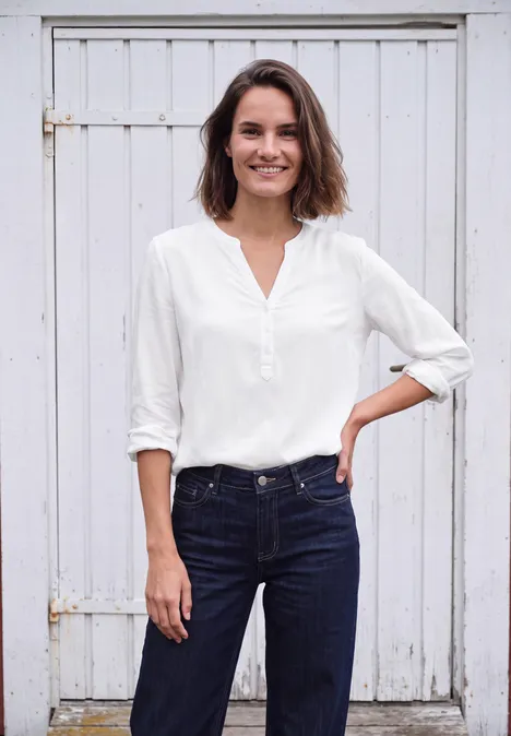 Frau in weißer Bluse und Jeans vor weißem Holztor, lächelt und hat die Hand in der Hüfte.