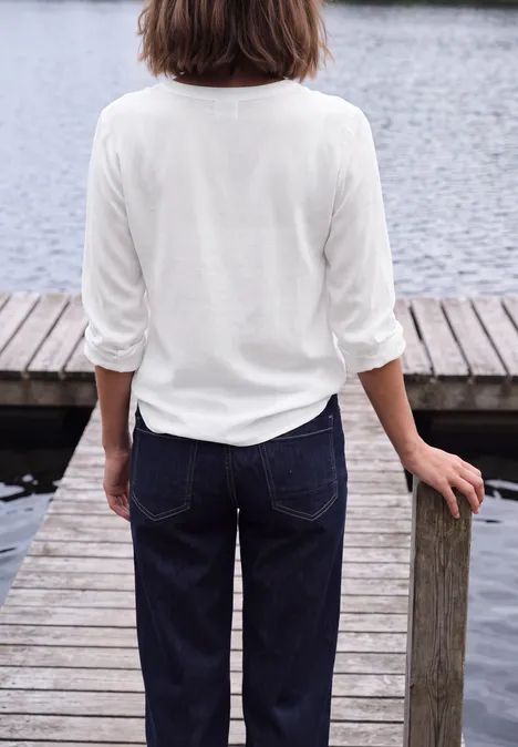Rückansicht einer Frau mit weißem Oberteil und Jeans auf einem Holzsteg am Wasser