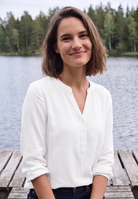 Junge Frau in weißer Bluse lächelt am Wasser, auf einem Holzsteg mit Wald im Hintergrund.