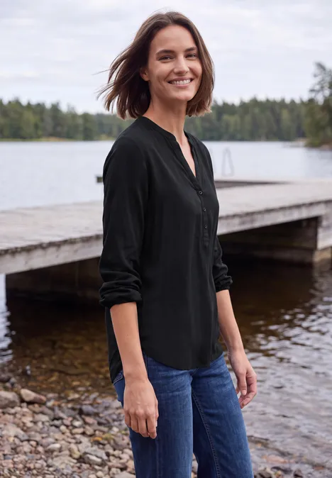 Eine Frau in schwarzer Bluse und Jeans steht lächelnd an einem Steg an einem ruhigen See.