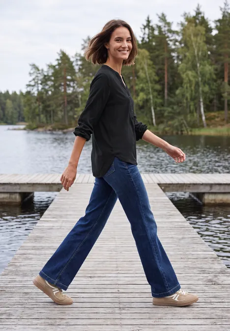 Eine lächelnde Frau in Jeans und Pullover spaziert über einen Steg am See, umgeben von Bäumen.