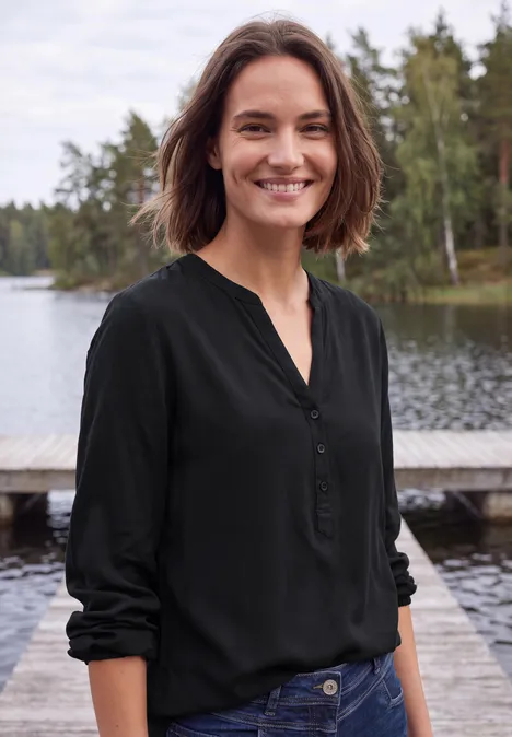 Eine lächelnde Frau in schwarzer Bluse steht an einem Steg am See.