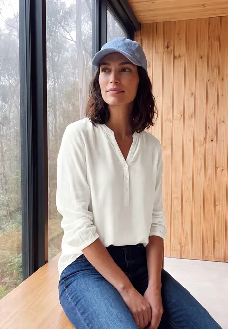 Femme décontractée en chemise blanche, jean et casquette, assise près d’une fenêtre en bois.