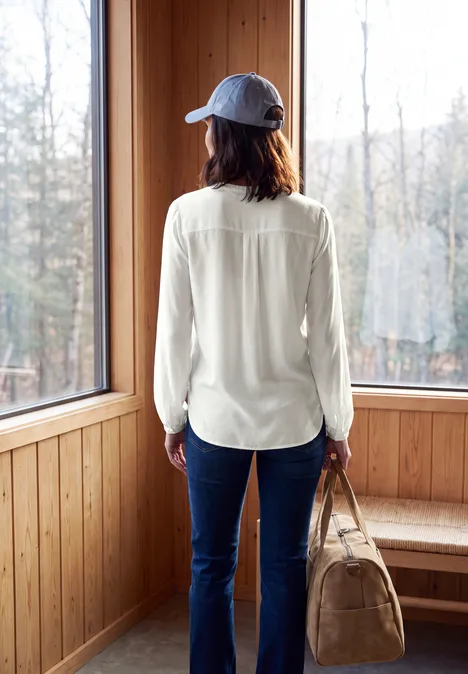 Femme en casquette, chemise blanche et jean, sac à la main, face à une grande fenêtre dans une pièce en bois.
