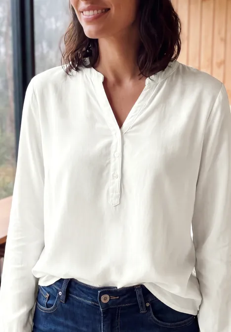 Femme souriante vêtue d'une blouse blanche près d'une fenêtre en bois