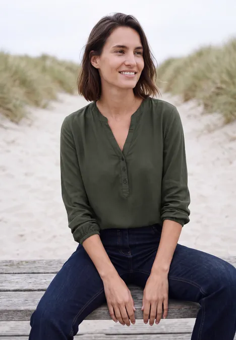 Eine lächelnde Frau sitzt entspannt auf einer Bank am Strand, trägt grüne Bluse und Jeans.