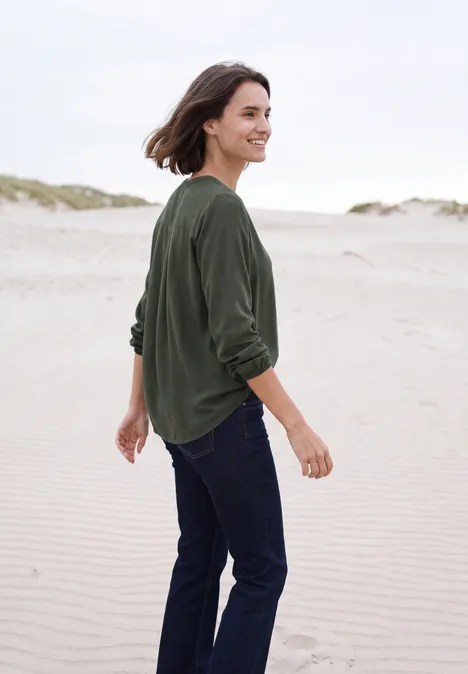 Eine Frau in grünem Pullover und Jeans spaziert am Strand.