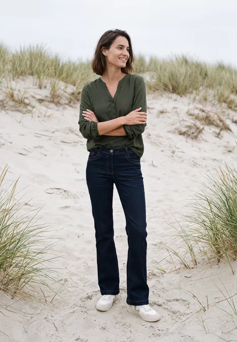 Frau in grüner Bluse und Jeans steht lächelnd mit verschränkten Armen am Dünenstrand.